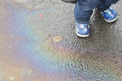 Child's feet wearing blue boots decorated with shark silhouettes next to a rainbow oil sheen on wet pavement.