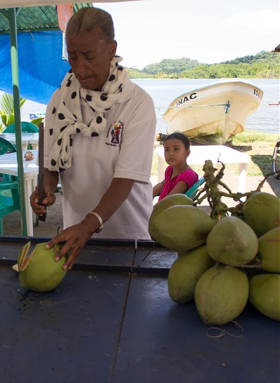 Beach vendor opening a green coconut with a large knife, a bunch of coconuts on the table nearby.