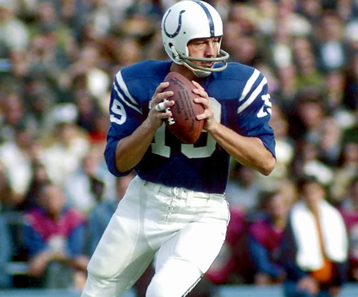 Foto do quarterback Johnny Unitas em jogo pelos Colts, em 1970. Ele está centralizado na imagem, com a arquibancada do estádio ao fundo, desfocada. Ele está vestindo uma jersey azul dos Colts, uma calça branca e um capacete branco com o símbolo do time em azul. Está fazendo o movimento para passar a bola.