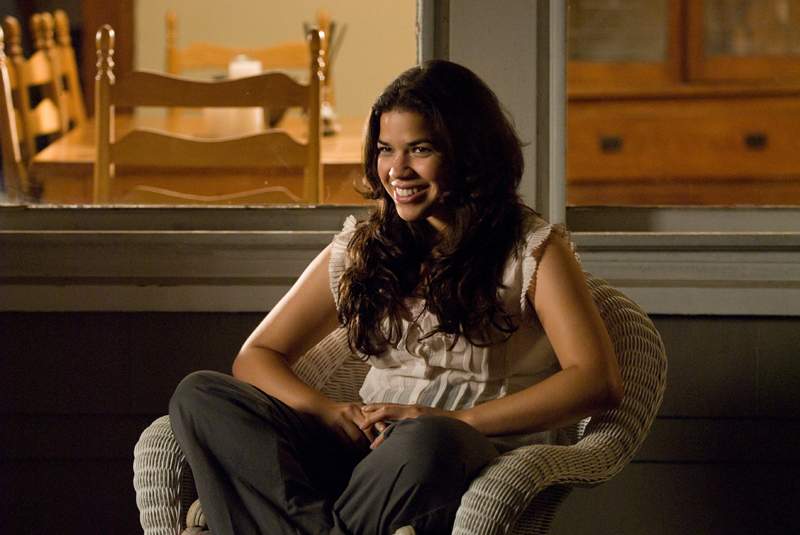 America Ferrera in Sisterhood of the Travelling Pants (2005). Carmen, a young Puerto Rican woman with long, dark curly hair, is sitting in a wicker chair out on a porch, wearing a white vest and grey trousers. She has her legs crossed underneath her and is smiling off-camera.