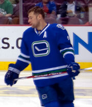 An ice hockey player, visible from the knees upwards, skating with spectators visible behind him. He is holding a hockey stick and wearing gloves and a sweater with a simple drawing of a hockey stick on the front, and a large "A" below his left shoulder. Not wearing a helmet, he is looking to his right, and has a relaxed expression.