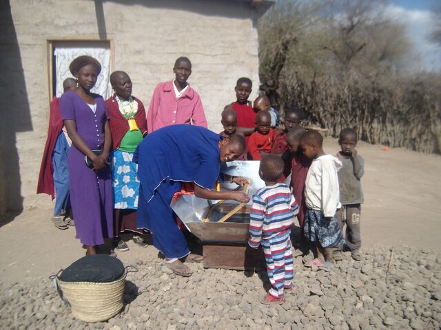File:Naomi removing food from solar cooker to fireless cooker-1.jpg